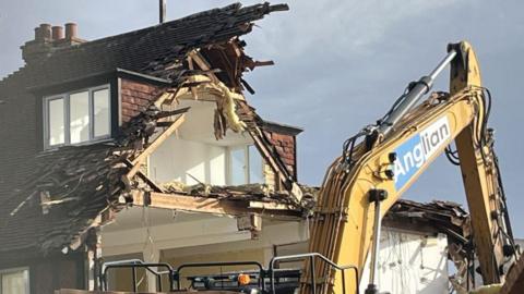 A large yellow crane in front of a partially demolished home. It is cloudy above.
