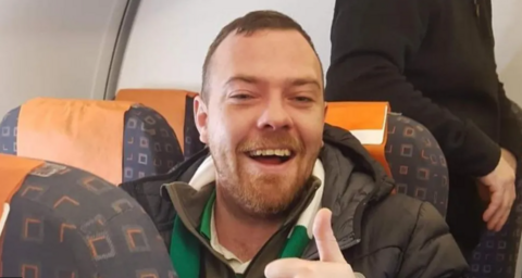 Kevin Davidson sitting down giving the thumbs up while wearing a green and white scarf and black coat