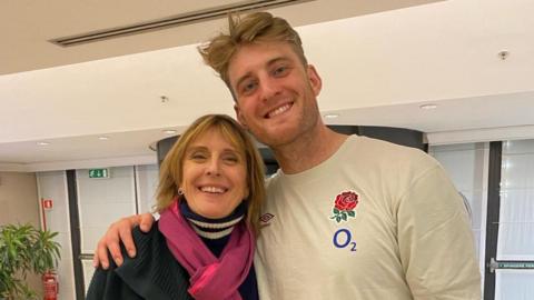 Alex Coles with his arm round his mother, the Reverend Olivia Coles. He is wearing a white T-shirt with the England red rose on the breast. She is wearing a blue jumper and a pink scarf. They are both looking at the camera and smiling.