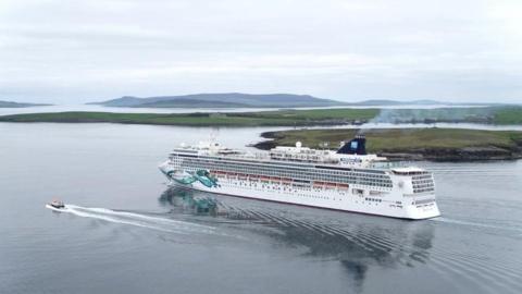 Large white cruise ship sailing past island scene with a support tug next to it, both making ripples in the water.