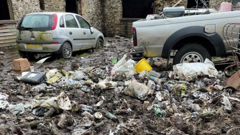 The image shows a large amount of discarded domestic waste with two vehicles including a small silver car which appears to have been abandoned and the rear of a pick-up truck containing more waste. There are stone farm buildings in the background which appear to be for animals.