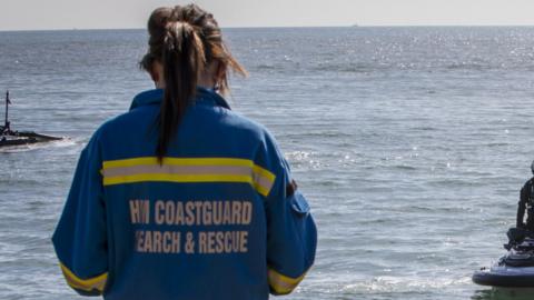 A coastguard rescue officer looks out to sea. The camera is behind them, so "HM Coastguard Search & Rescue" on the back of their uniform is visible.