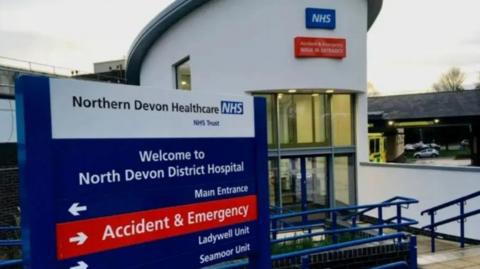 A signpost saying "Northern Devon Healthcare NHS Trust" and reading "Welcome to North Devon District Hospital". Under that are arrows pointing to the main entrance, accident and emergency, the Ladywell Unit and the Seamoor Unit. 