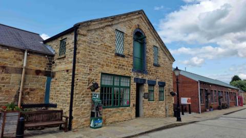The exterior of the visitor centre. It is built of stone and is two storeys high. It sits on a stone paved area, and old black lamp post is next to it