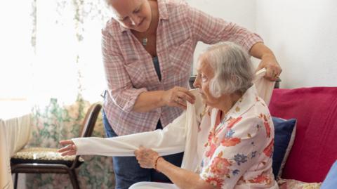 A woman helps another older woman sitting on a sofa put on her cardigan. 