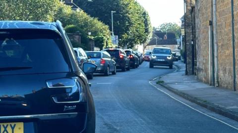 A line of stationary traffic viewed from behind on a gently curving road with hedges on one side and sandstone buildings on the other