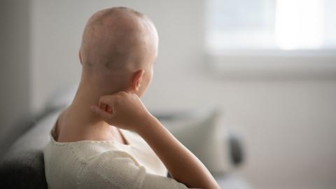 A woman is indoors in her living room. Her head is shaved due to chemotherapy. She is sitting and looking thoughtful but you can't see her face
