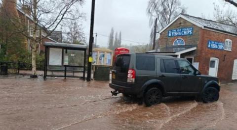 A dark green Land Rover is driven through floodwaters in Ewyas Harold, Herefordshire, in the background there is a bus stop and a fish and chips shop.