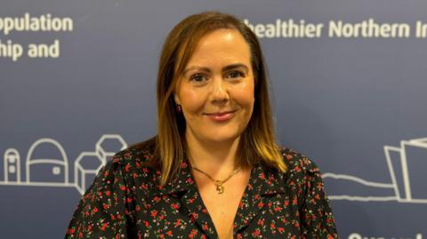 A close-up image of Rachel Spiers standing in front of a dark background with some text written in white. She is a young woman with straight, brown hair at shoulder length and she is wearing a floral patterned dress and a gold necklace. She is smiling at the camera with her mouth closed.