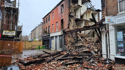 A collapsed building at the centre of a terrace of three storey buildings with shops at street level. All that is left of the collapsed section is a blackened steel beam that stretches across the top of the former shop front. A huge pile of bricks and charred wood spills out across the road.