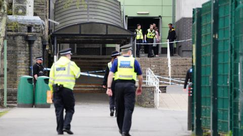 A police cordon at the school. Two officers are walking away from the camera, one wearing a fluorescent jacket and hat and the second wearing a fluorescent vest. More officers can be seen at the top of a flight of stairs by the school entrance. There is police tape across the top of the stairs, and also at the entrance to covered walkway to the left hand side. 