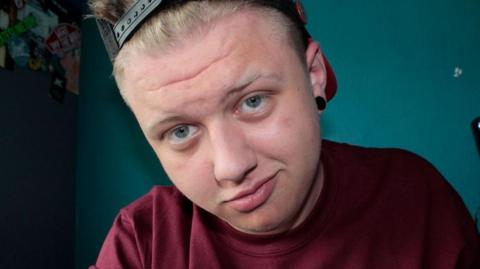 Liam Andrews is wearing a maroon T-shirt and back-to-front, black baseball cap and is staring right into the camera with a quirky slight smile.