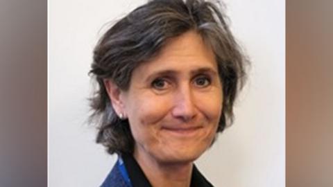 A woman with a brown/grey bob wearing a blue jacket smiles at the camera side on.