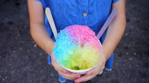 A child wearing a blue dress holding a multi-coloured slushy ice drink out in front of them with both hands around it.