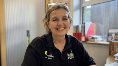 Sarah Comish has short blonde hair and is wearing a black polo shirt with the Manx NFU logo on the front. She's smiling, sitting in her office with a window to the right and a storage unit behind her on the left.