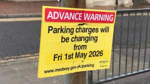 A yellow sign attached to a railing. It reads "Advance Warning: Parking charges will be changing from Fri 1st May 2026"