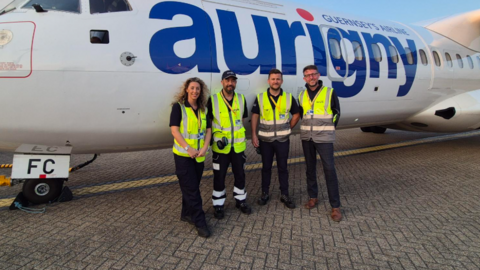 Four people dressed in black clothes and green high visibility vests. They are stood in front of the side of a Aurigny plane. 