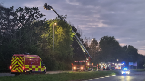 Multiple fire engines and an aerial ladder platform block part of a dual carriageway at dusk. The platform is extended over a row of trees which are obscuring a house that is on fire.