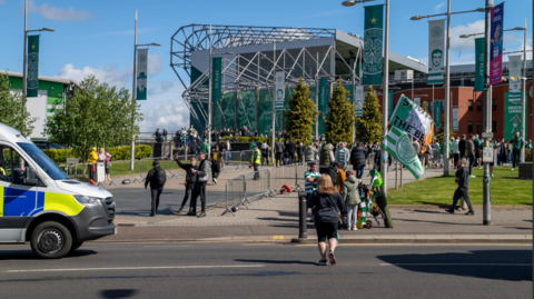 Football fans arrive at Celtic Park in Glasgow. The stadium can be seen in the background. A police van is parked on the street. 