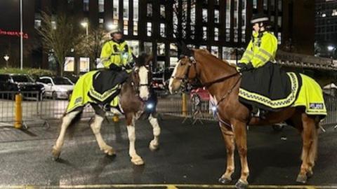 Two police officers riding police horses during the evening, in front of a block of flats. The horses, one is mainly brown, the other brown and white, and officers are dressed in hi-vis jackets.