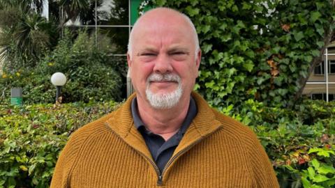 Andy Gealer is wearing a mustard coloured fleece stands in front of some bushes and is looking into the camera