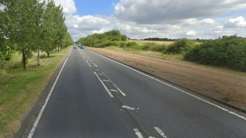 Dead straight road labelled 'A130' by Google with young trees on the left hand side of the road and farmland behind hedges on the right hand side. 