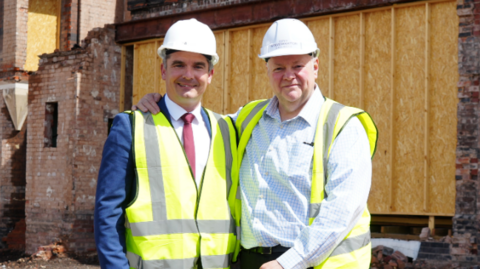 The two men men stand on an area of brown soil with one a spade. Dr Cross is on the left with a blue suit and brown shoes and Mr Simkins has dark trousers and a blue shirt on. Both have florescent jackets on and hard hats. Behind them is a boarded up building  and another redundant brick building in the distance.