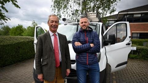 Nigel Farage, in his 60s, wearing a tweed jacket, a chequered shirt and red tie, smiles at the camera standing next to Robert Kenyon, wearing a blue bubble coat and jeans, who is also smiling and has his arms folded. They are standing on a driveway in front of a white van with ladders fixed to its roof. 