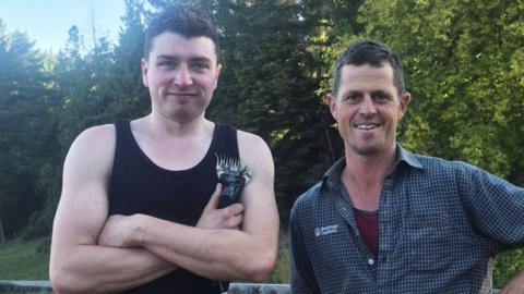 Two men with dark hair, one with a black vest and holding shearing clippers and the other with a blue checked shirt in front of trees.