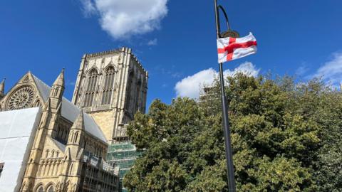 An England flag sits on a lamppost against the backdrop of York Minster. Some trees are also in the background.