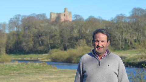 Northumberland County Councillor Patrick Lambert. He has short salt and pepper hair and is wearing a grey jumper. He is standing in an open area surrounded by grass, with a stream and a castle ruin in the background.