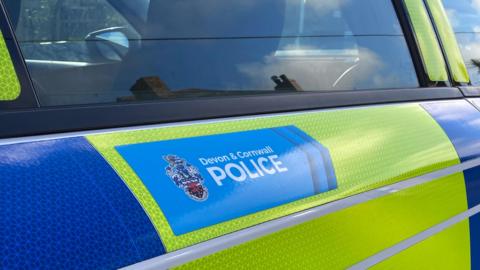 A close up of the door of a police car which has the Devon and Cornwall Police lettering and a shield on a badge. The car is liveried in fluorescent yellow and blue. 