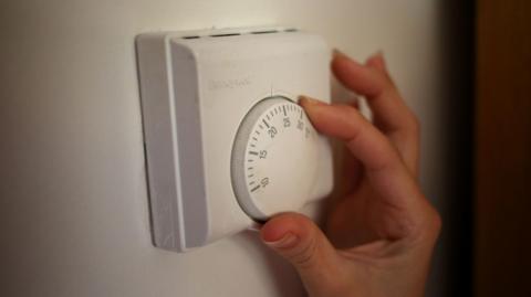 A hand adjusts a heating thermostat. The unit is a white square with a dial in the middle.