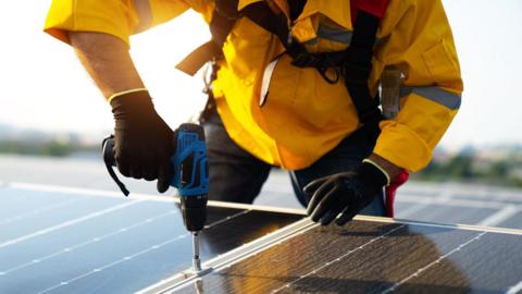 Worker wearing a hi-vid jacket installing a photovoltaic solar panels on a roof. He is using a blue drill with a screwdriver attachment.
