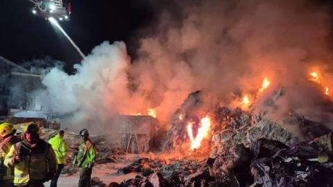 The scene of the fire at night. A small cluster of firefighters are on the left of the photo in high-vis clothing and helmets, with an aerial platform at top left squirting water on to the fire below. Flames are leaping from the mounds of rubbish or recycling with large plumes of smoke above