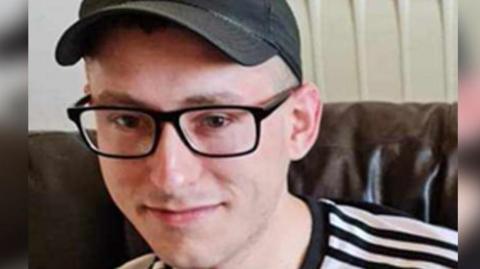 Martyn Richardson, wearing glasses and a cap, looking at the camera. He is sitting on a sofa and is wearing a black and white top.