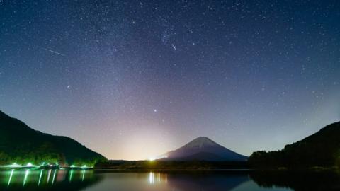 a snow capped mountain of Mount Fuji, green and yellow light reflecting onto a lake under a blue and pink starry sky with white streaks and dots or a meteor shower
