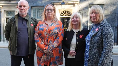 Steve Kelly, who has a bald head, white stubble and a green jacket, Charlotte Hennessy, with light ginger hair and wearing an orange floral dress, Margaret Aspinall, with shoulder length blonde hair and a black jacket, and Sue Roberts, who has blonde hair and a grey and black woollen coat, all stand looking into the camera with determined expressions with the black door of Number 10 Downing Street in the backdrop.