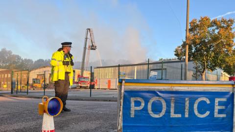 A police officer standing in front of a warehouse at an industrial estate following an explosion and fire. The officer is wearing a black police hat and a high vis jacket. There is a blue police sign on the ground. Smoke can be seen in the background as well as equipment from the fire crews.