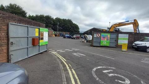 Gated entrance to a household recycling centre. The metal gates are open and cars are parked inside. The arm of a yellow digger is poking above the brick wall next to the gate.