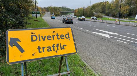 A yellow sign diverting traffic off the central motorway in Newcastle. Two cars are leaving the road and heading up a slip road. Rose Bridge can be seen in the distance across the motorway.