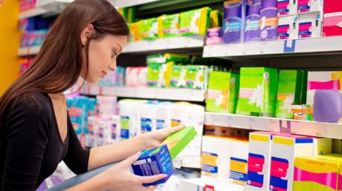 A young woman, wearing a black top and jeans, crouches down to look at period products that are stacked on a low shelf in a shop. She has selected two boxes that she examines in her hands.