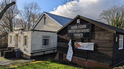 Riddlesden United Reformed Church on Scott Lane, with a white building on the left and a wooden hut on the right. Bare trees are behind, with blue sky and clouds. Green grass is in front.