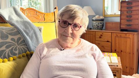 Ann Campbell is an older woman. She is short white hair and is wearing glasses and a light pink jumper. Her room is furnished with a wooden dresser and cushions.