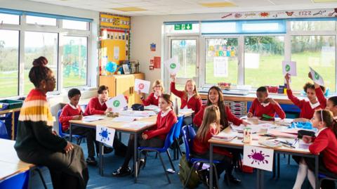 A teacher addresses a group of children at desks in a classroom