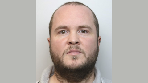 A custody picture of Matthew Ely. He has a dark scruffy beard and thinning dark brown hair on his head. He has small silver-coloured piercings in his left ear and just below his mouth