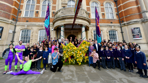 Dancers, performers, local dignitaries and schoolchildren gather outside the entrance to Battersea Arts Centre in front of silk banners and a yellow floral representation of the borough's new logo (a stylised interpretation of the borough's boundaries made to look like the letter w)