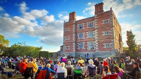 A large block-shaped brick building with four floors and large brick chimneys with a crowd of people sitting outside on camp chairs looking toward the building
