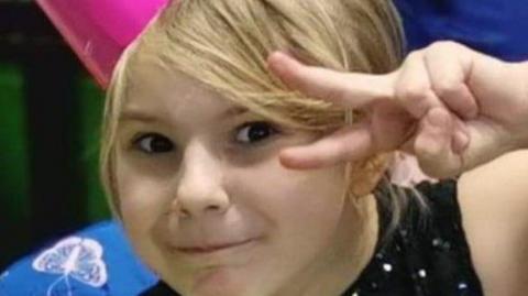 A young girl with blonde hair at a birthday party is posing for the camera. She has a pink party hat on and is making a peace gesture with her fingers in a v-sign.