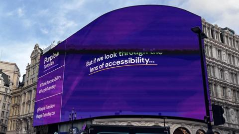 A large digital display board in Piccadilly Circus displays a message about Purple Tuesday. 